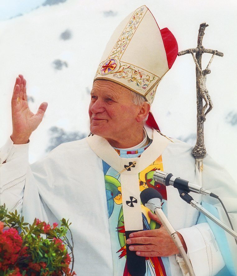 Pope St. John Paul II in white vestments and mitre, arms raised in greeting at an outdoor Mass.