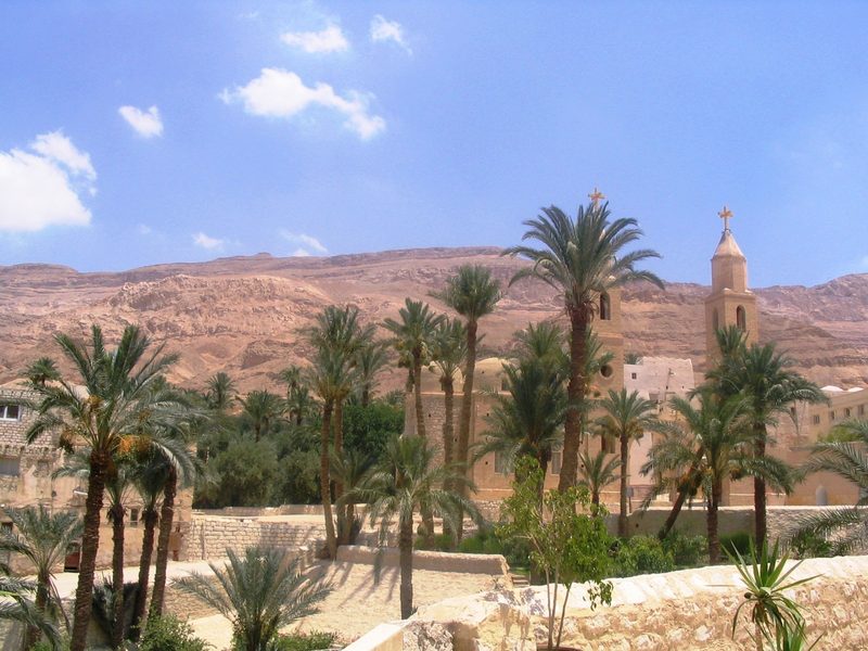 The Monastery of St. Anthony in the Egyptian desert: palm trees and pale buildings at the foot of bare red mountains.