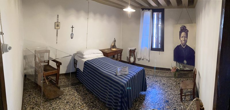 The simple room of St. Josephine Bakhita at Schio: a narrow bed with a blue cover, a chair and a crucifix.