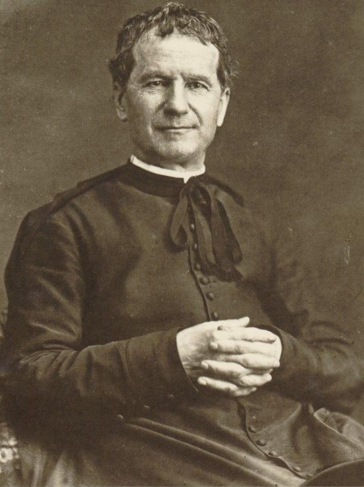 A photograph of Don Bosco in Turin in 1880: an elderly priest in a cassock, seated, with a gentle face.