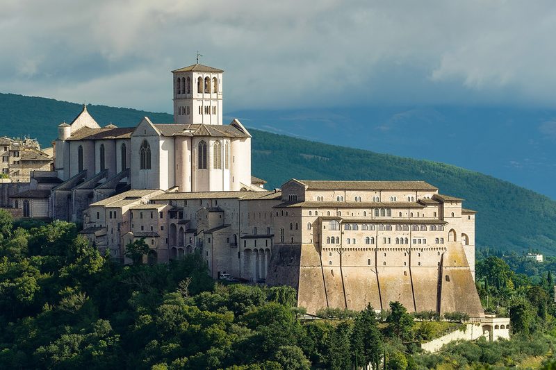 The Basilica of San Francesco in Assisi, seen from the hillside: two stacked churches and a long arcade above the Umbrian plain.