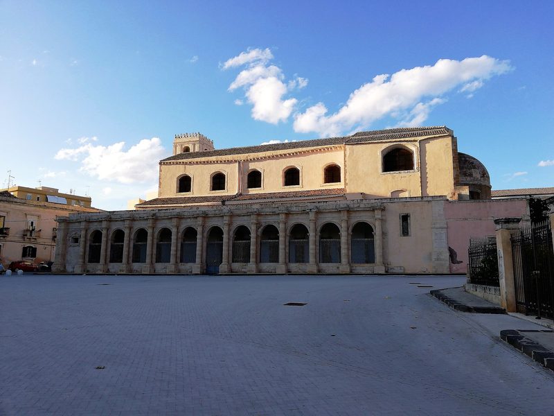 The church and arcaded buildings of Santa Lucia al Sepolcro in Syracuse, Sicily, under a blue sky.