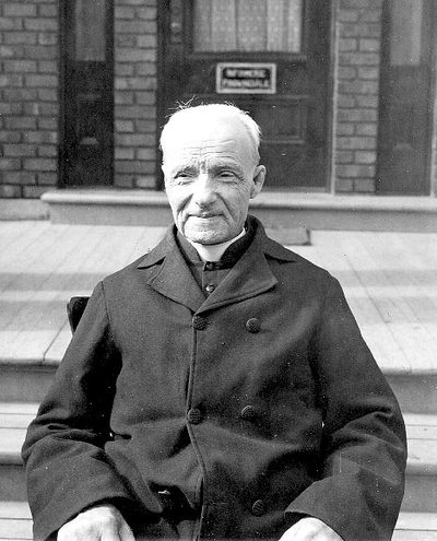 Black-and-white photograph of St. André Bessette, white-haired, seated in a dark cassock.