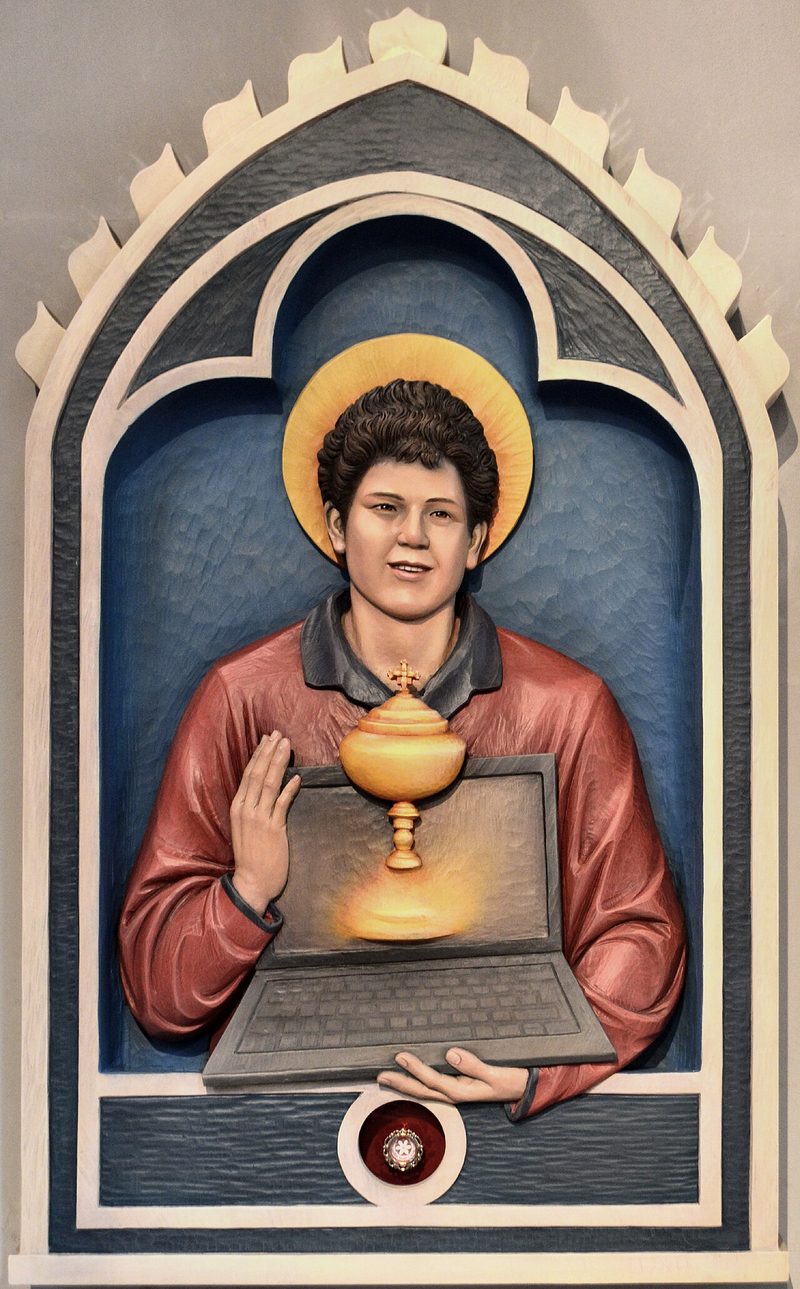 Painted carving of Carlo Acutis with a halo, holding a monstrance, in a church memorial.
