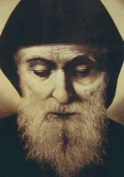 Photograph-like portrait of St. Charbel Makhlouf with a hooded monk’s habit and long white beard, eyes lowered.