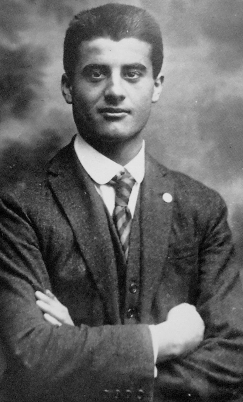 Black-and-white photograph of St. Pier Giorgio Frassati in a suit and tie, arms folded.
