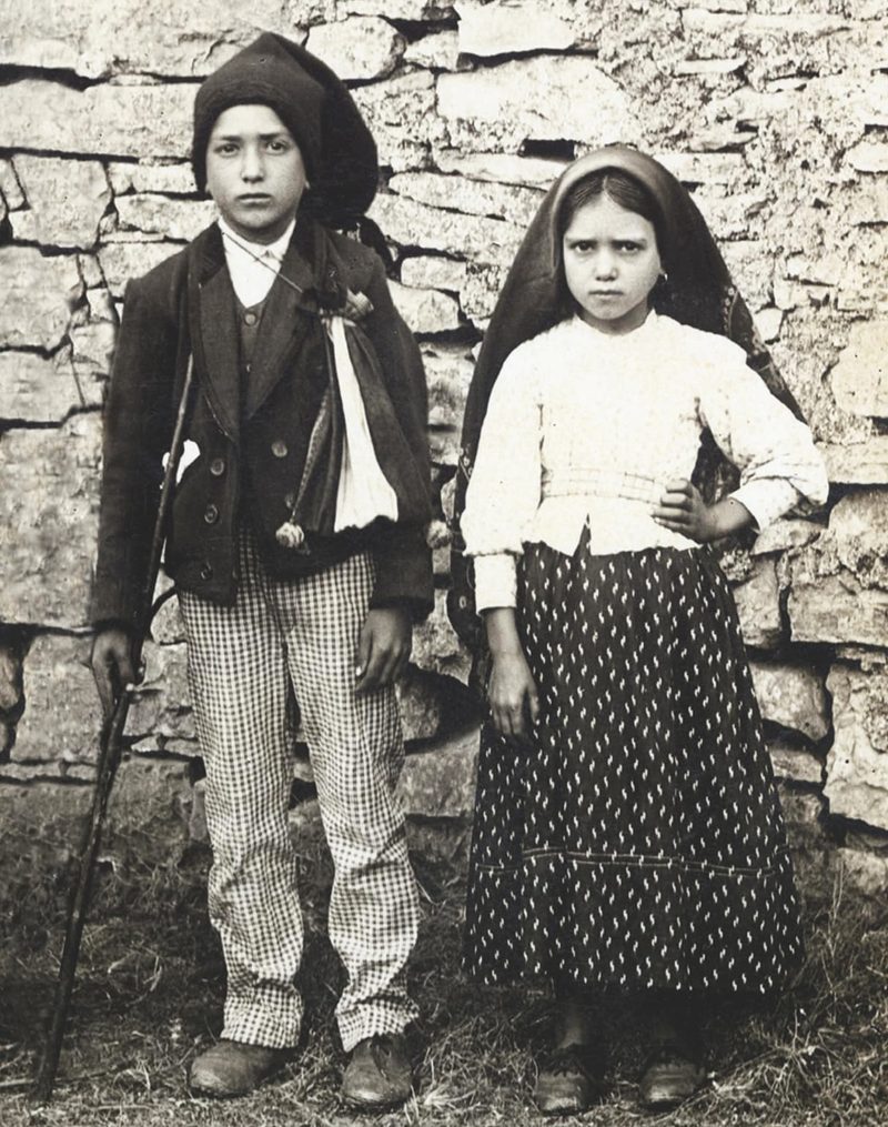 Black-and-white photograph of Francisco and Jacinta Marto standing by a stone wall.