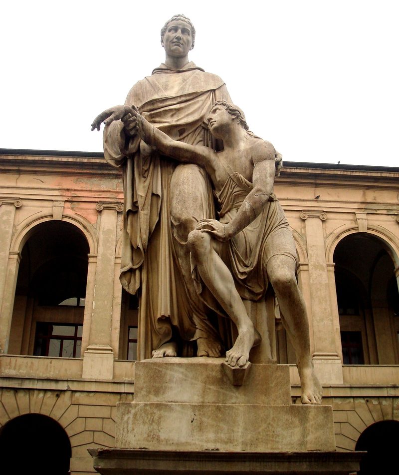 Marble statue of St. John of God carrying a sick man, in front of an arcaded building.