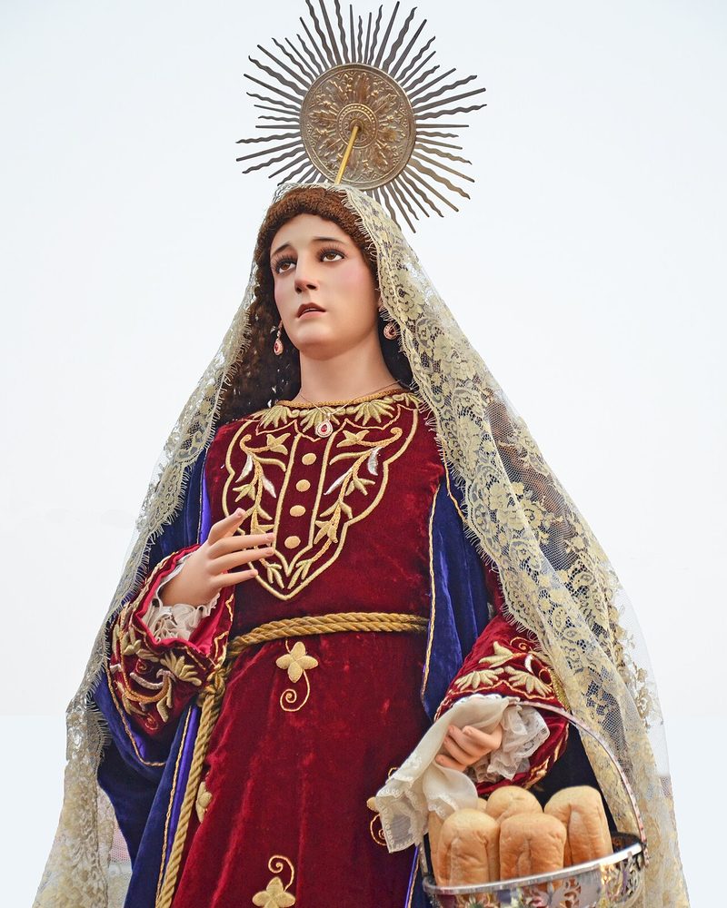 Processional statue of St. Martha in a red gown, crowned with a halo of rays, holding a basket of bread.